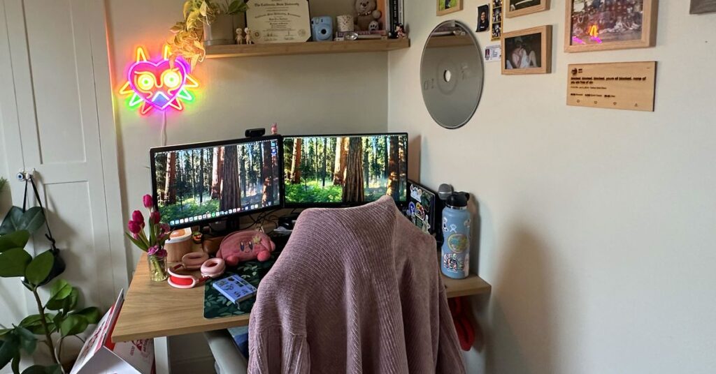 Desk in a corner with monitors, photos on the wall, and a chair with a sweater draped over it.