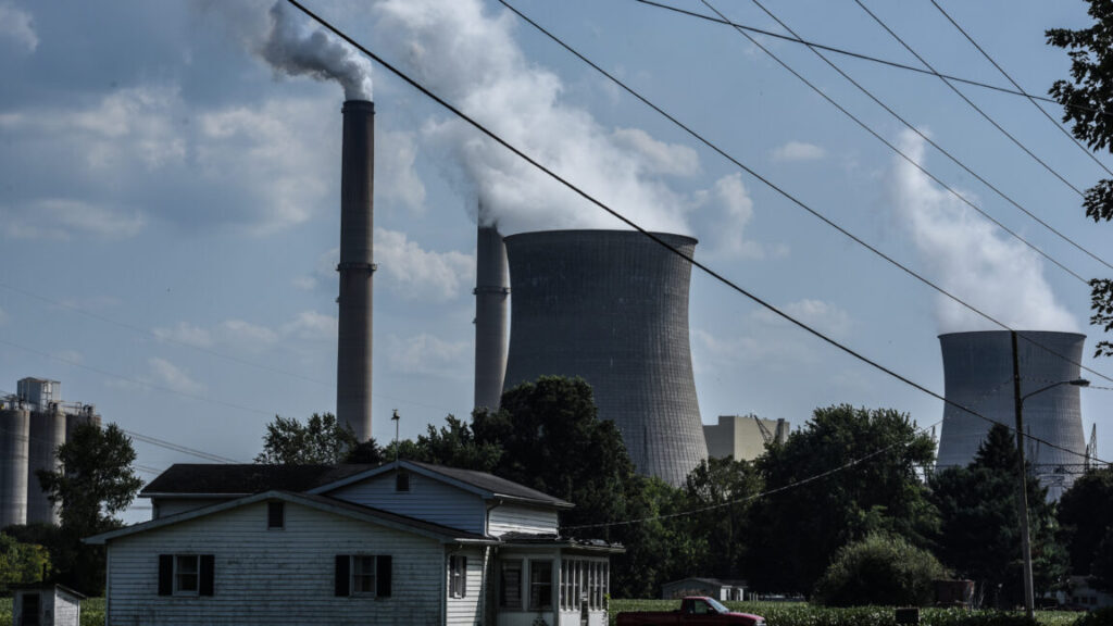 A house is in the foreground of two smokestacks that are emitting plumes.