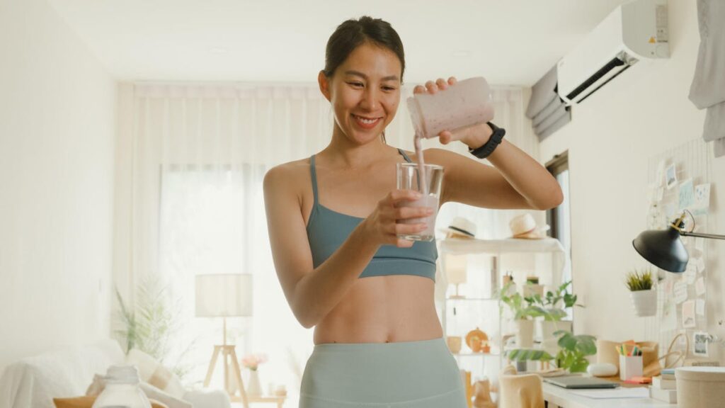 A person with pulled-back dark brown hair and a medium-blue sports bra pouring a smoothie into a glass.