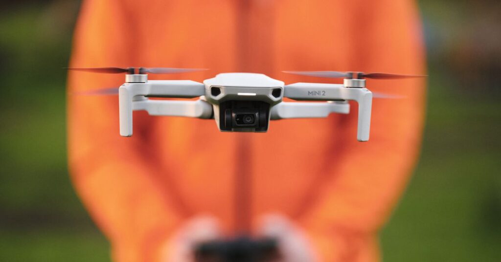 A drone faces the camera, in flight, with a blurred person in the background shoulders down in an orange puffy jacket controlling that drone, serving as an orange background.