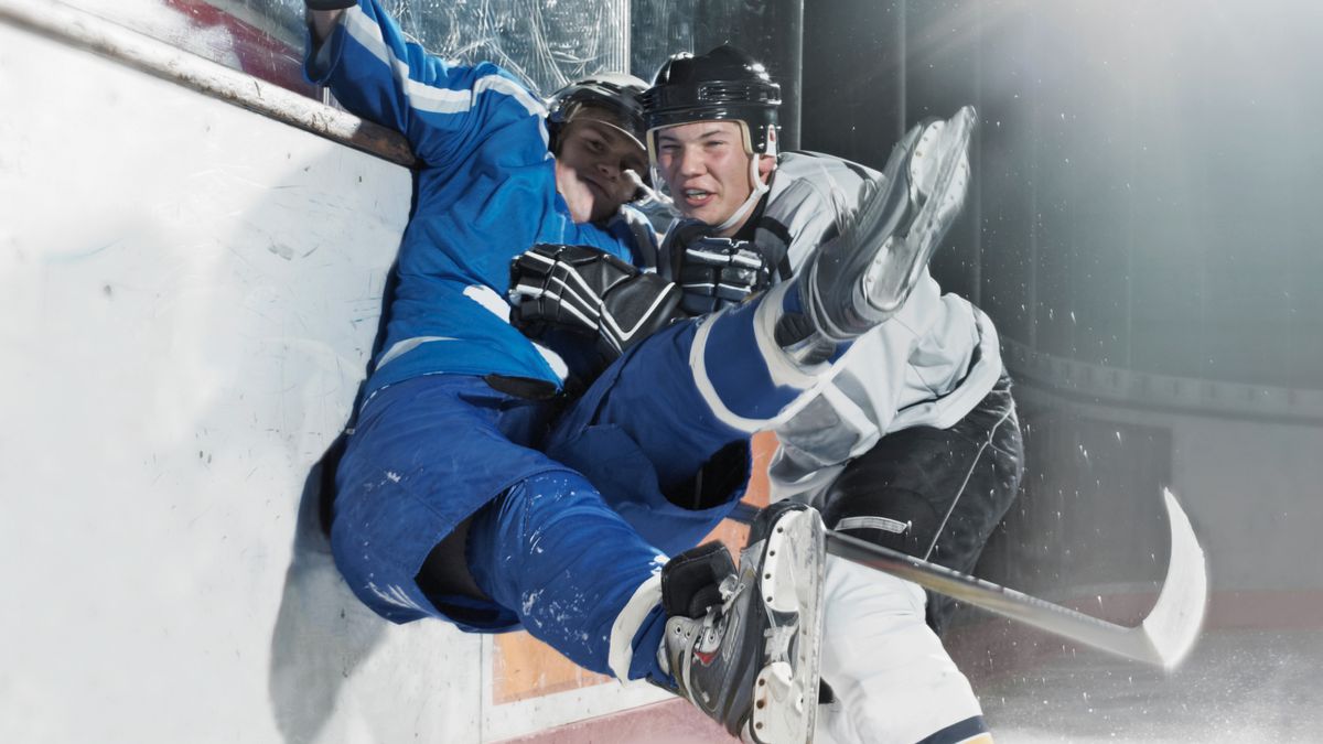 an ice hockey player in a white uniform slamming a second player in a blue uniform up against the wall of a rink