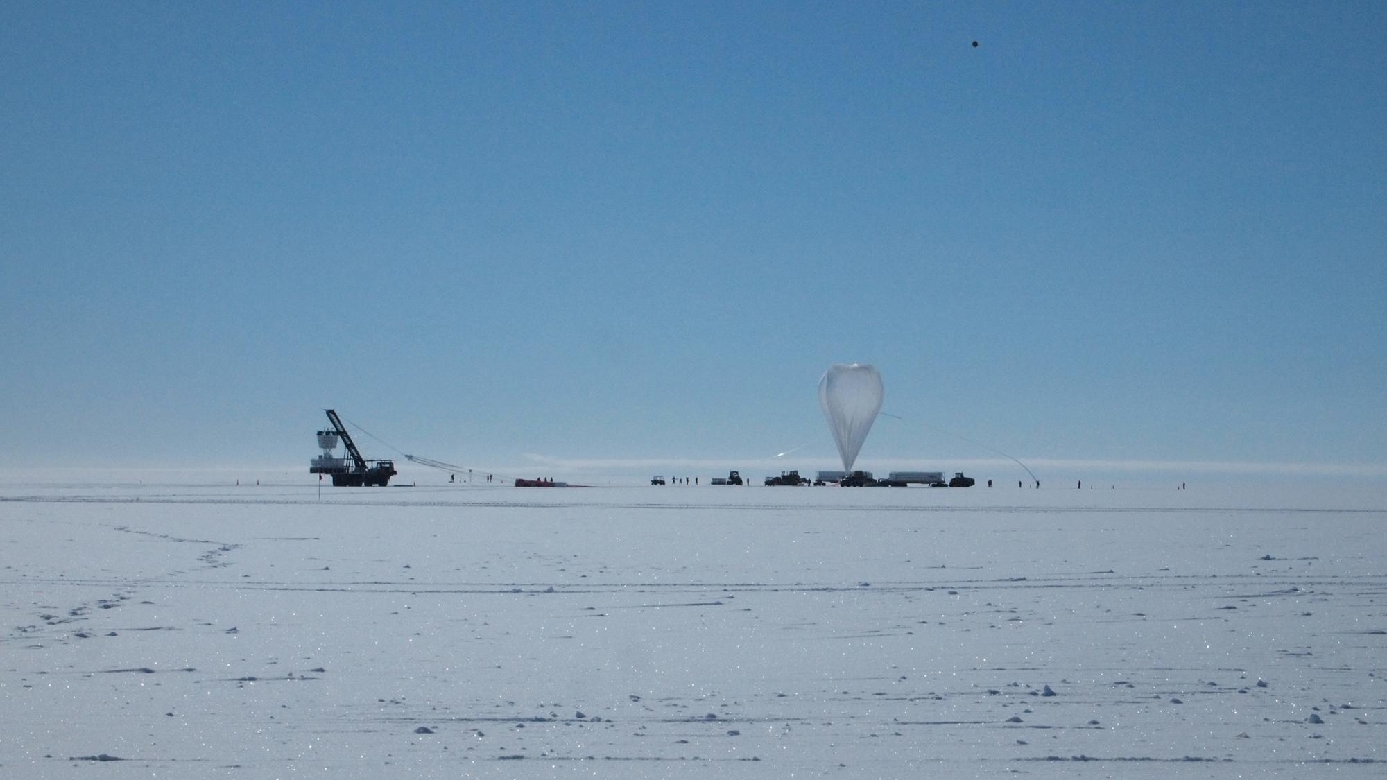 A balloon and science equipment sit on the surface of ice against the backdrop of a clear blue sky.